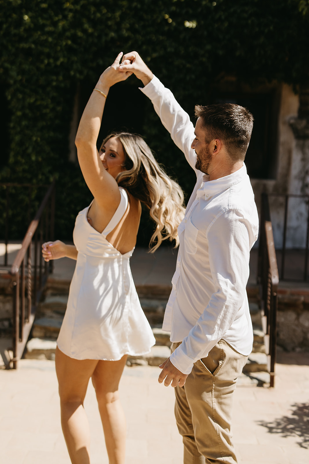 A couple stands close and embraces on a sunlit patio, with steps and a dark doorway in the background for their candid engagement photography 