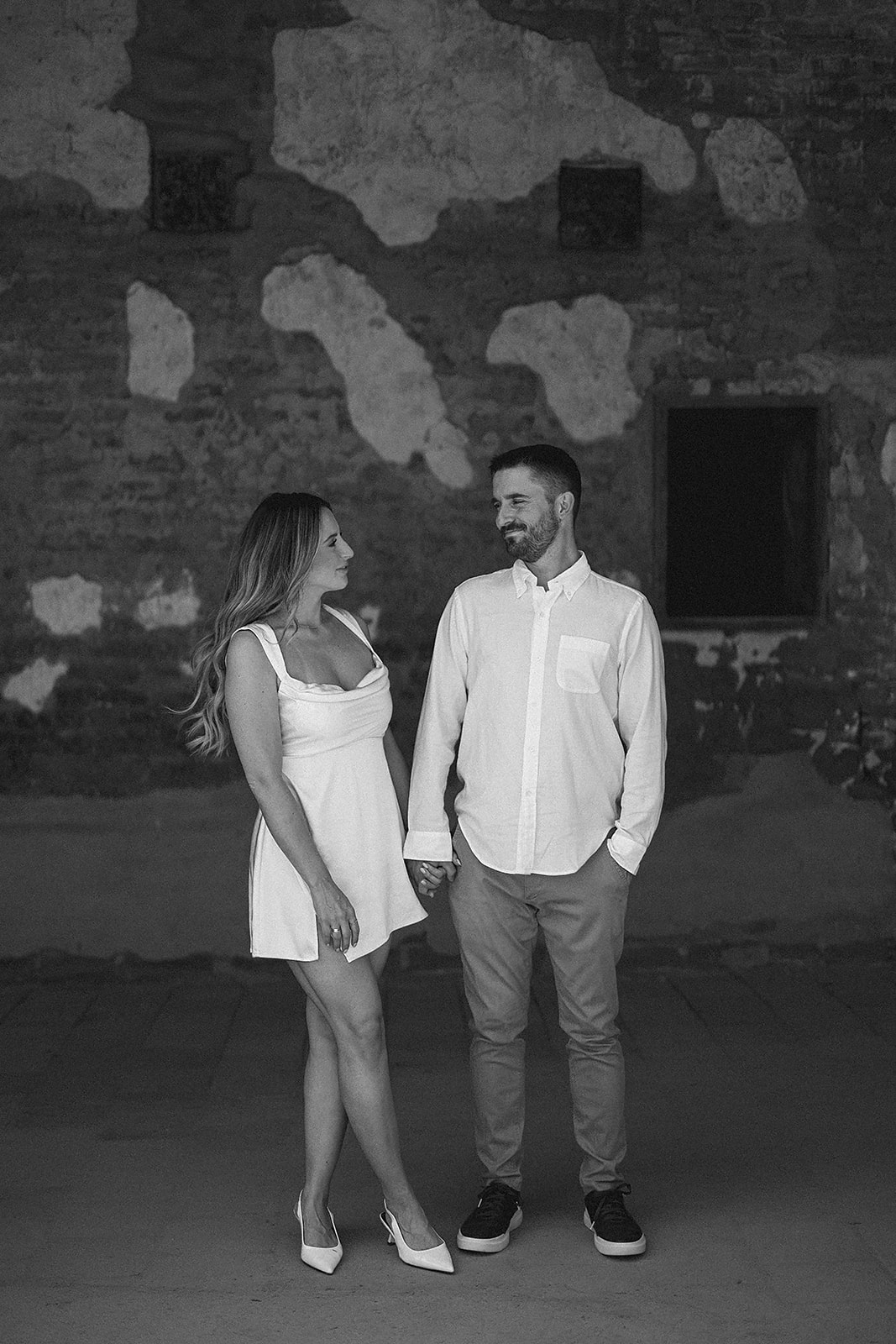 A couple stands holding hands against a textured wall. She is in a white dress, and he is in a white shirt and pants. Black and white photo.