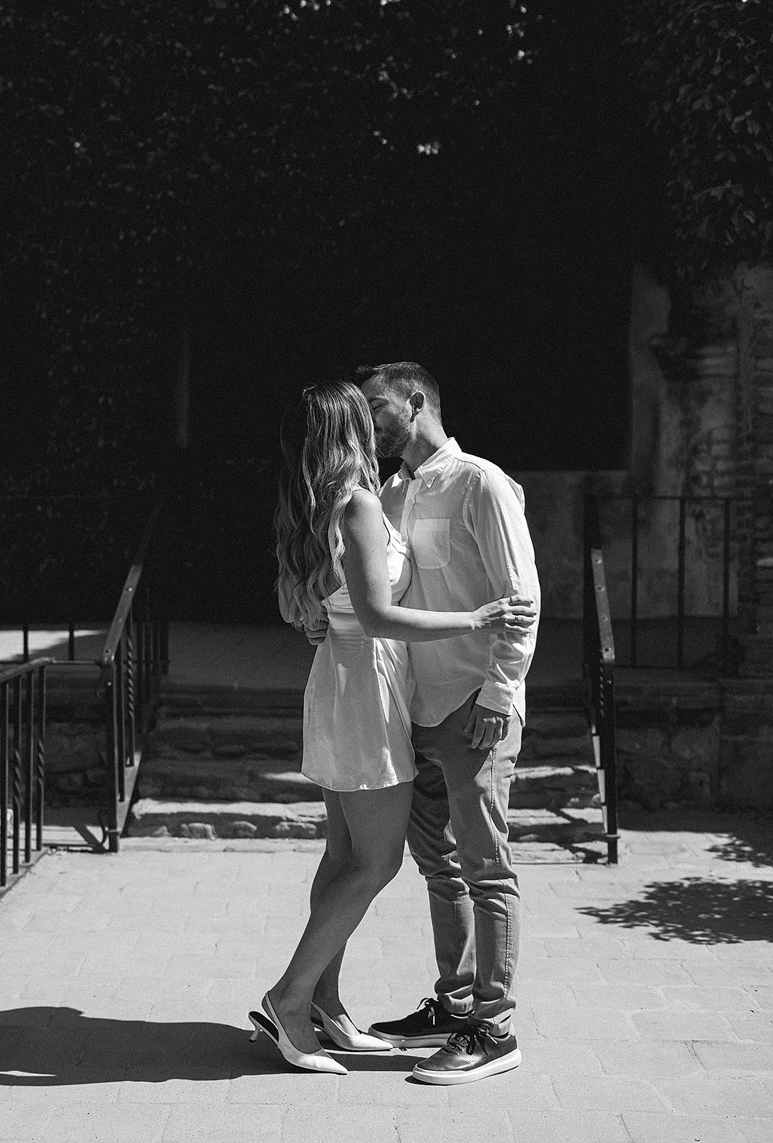 A couple stands close and embraces on a sunlit patio, with steps and a dark doorway in the background for their candid engagement photography 