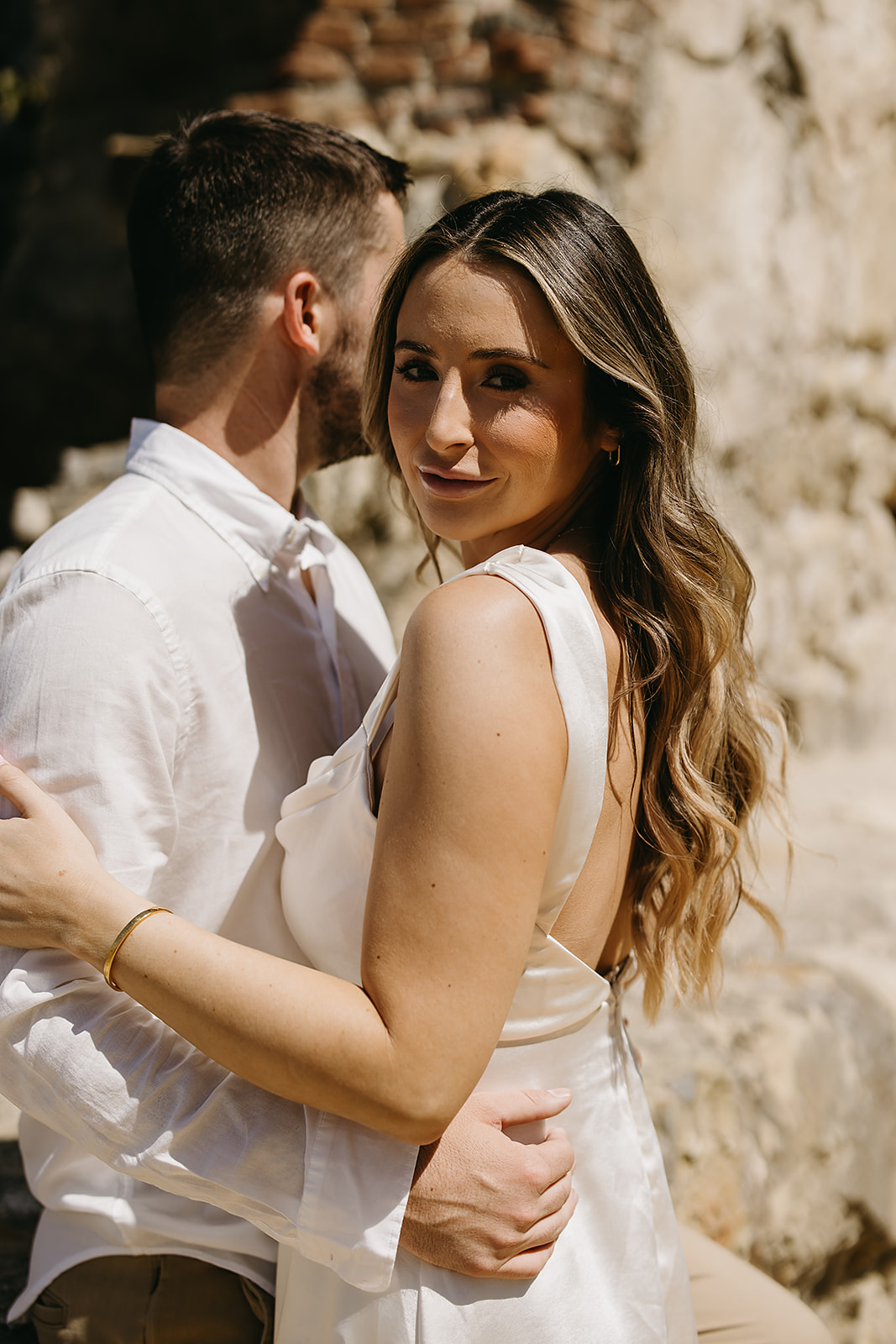 A couple in formal attire embraces outdoors, with the woman looking at the camera and the man facing away.