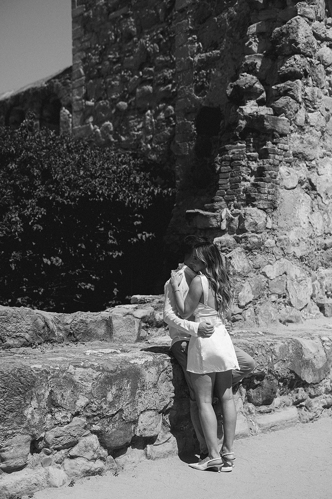 A couple embraces while kissing against an old stone wall on a sunny day.