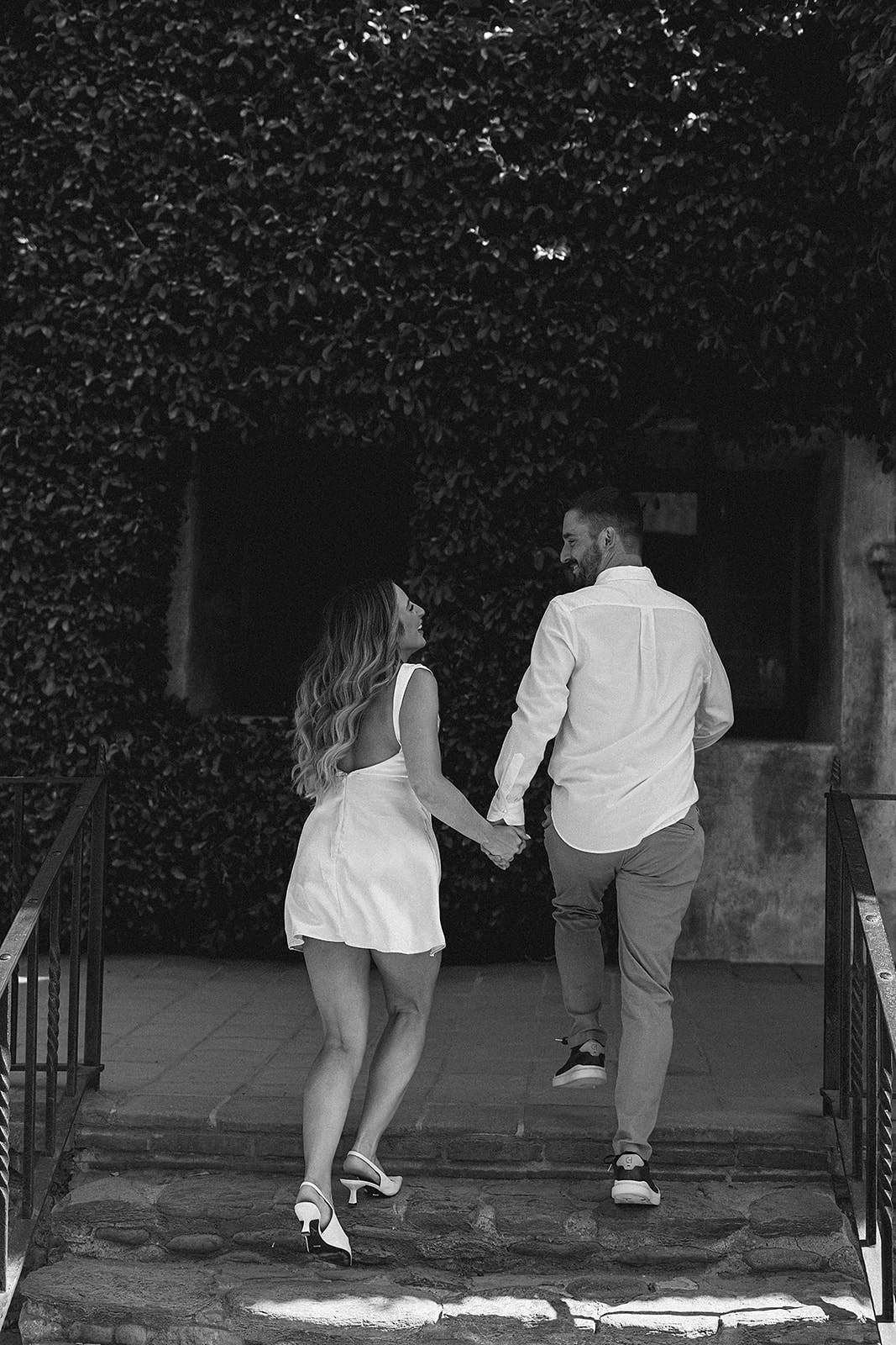 A couple stands close and embraces on a sunlit patio, with steps and a dark doorway in the background for their candid engagement photography 