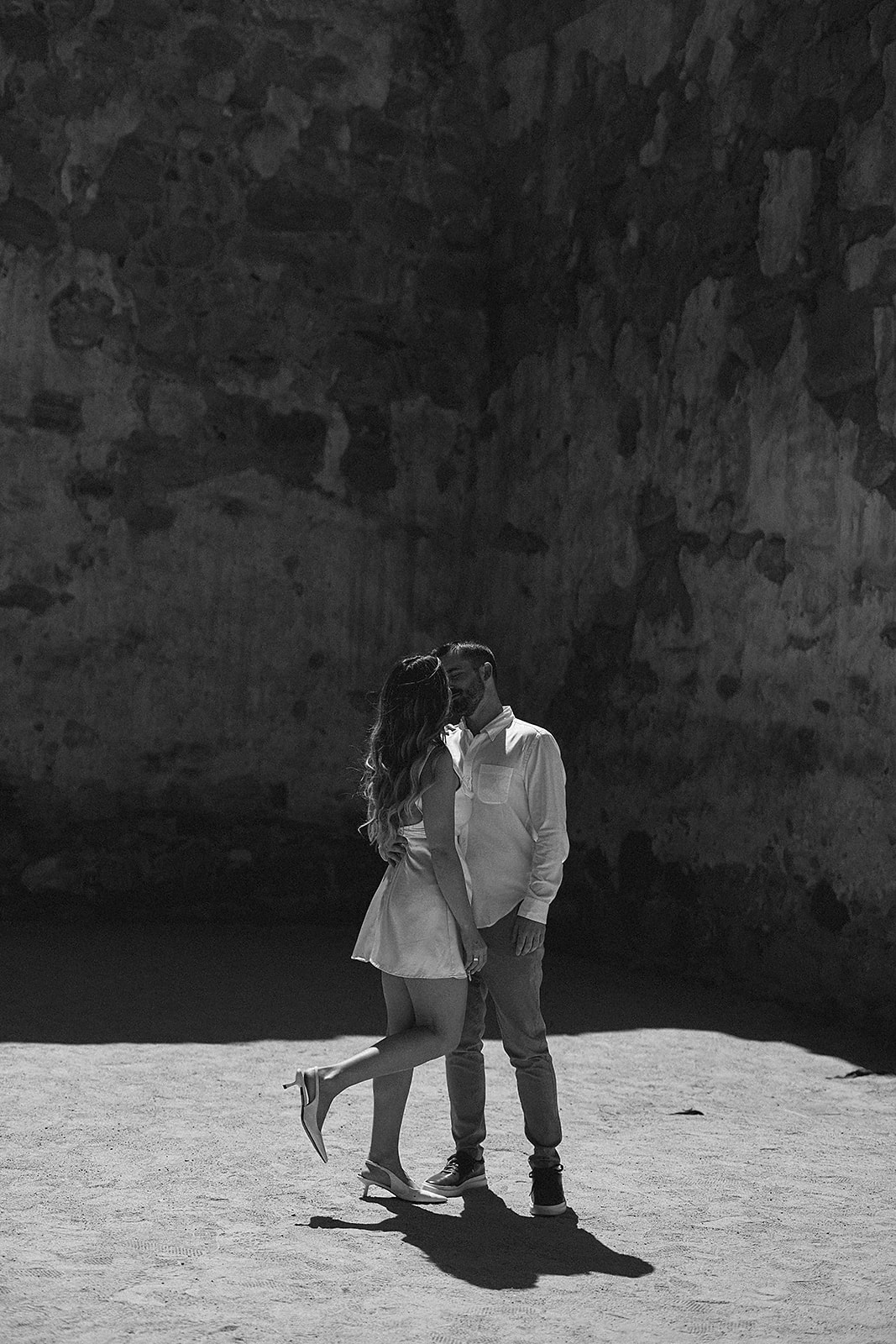 A couple kisses in a shadowed, textured stone-walled area under soft sunlight for their candid engagement photography 