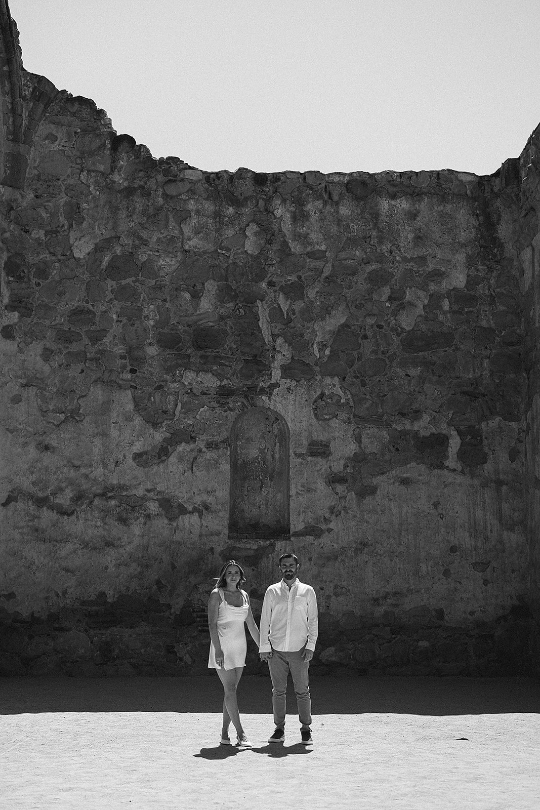 A couple kisses in a shadowed, textured stone-walled area under soft sunlight for their candid engagement photography 