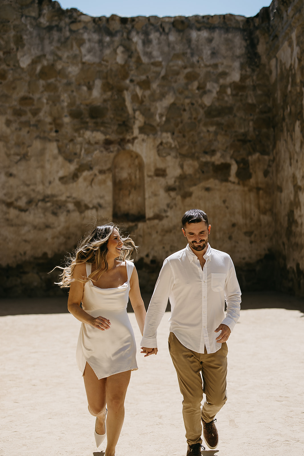 A couple in white clothing holding hands and walking in a sunny open space with stone walls.