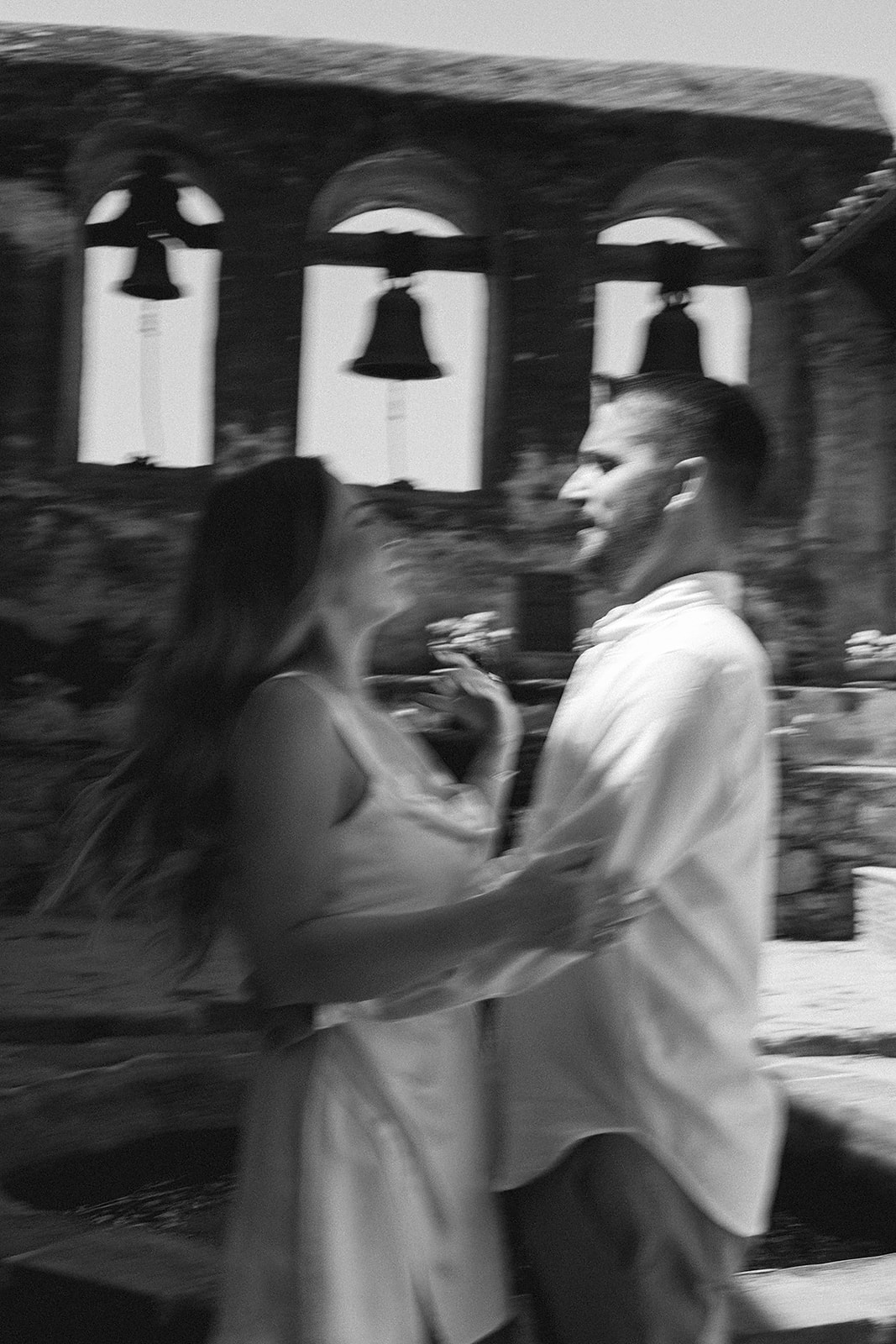 A blurred black and white image of a couple dancing outdoors in front of a historic building with three bells.