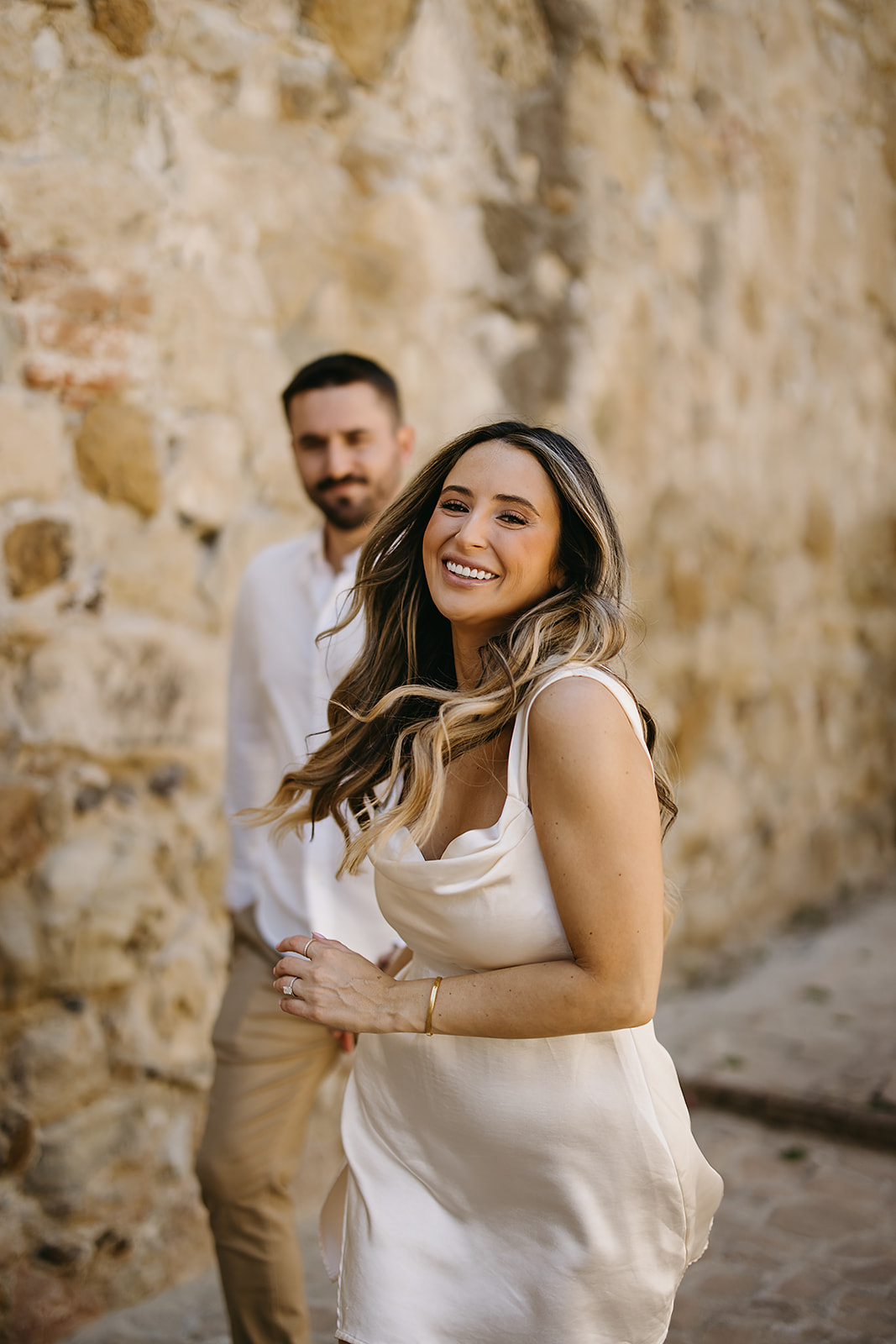 Woman in a white dress smiles while walking beside a man in front of a stone wall.