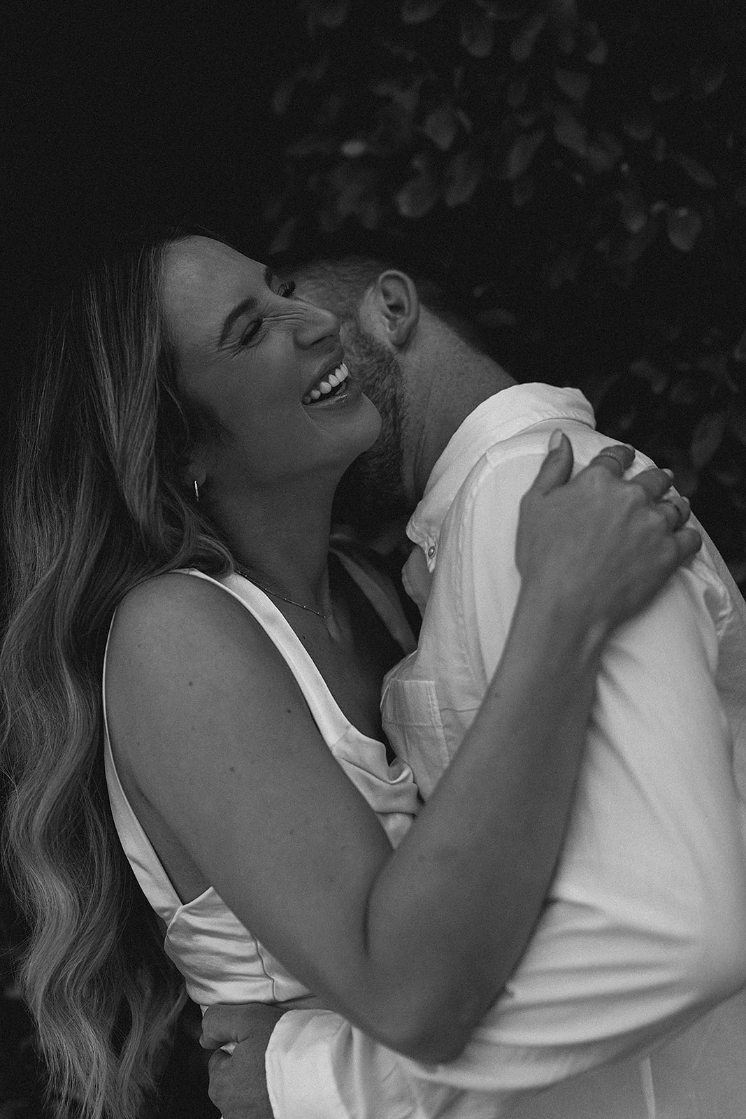 A couple embracing and smiling, standing close together. The woman has long hair and is wearing a sleeveless top, while the man is in a long-sleeve shirt for their candid engagement photography. The image is in black and white.