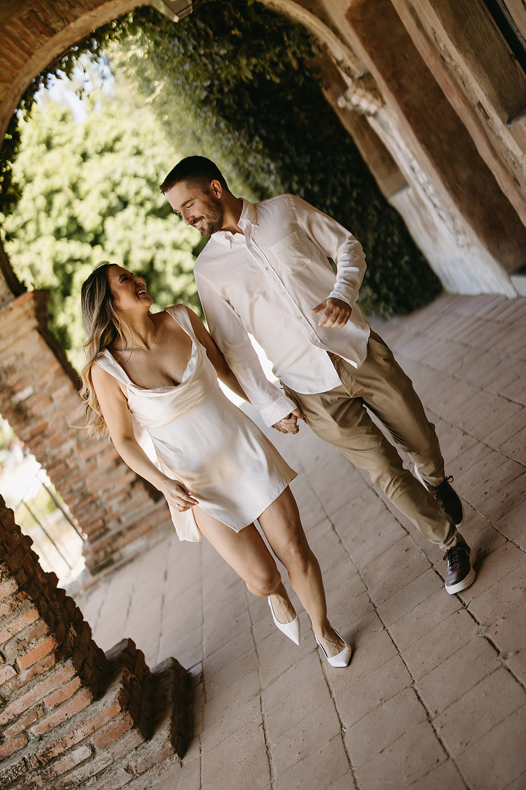 A couple walks hand in hand under a brick archway, smiling at each other. The woman wears a white dress, and the man wears a white shirt and khaki pants. They appear happy and relaxed for their candid engagement photography 