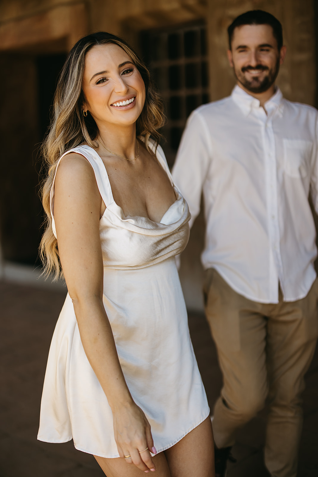 A woman in a white dress smiles at the camera, while a man in a white shirt and brown pants stands in the background.