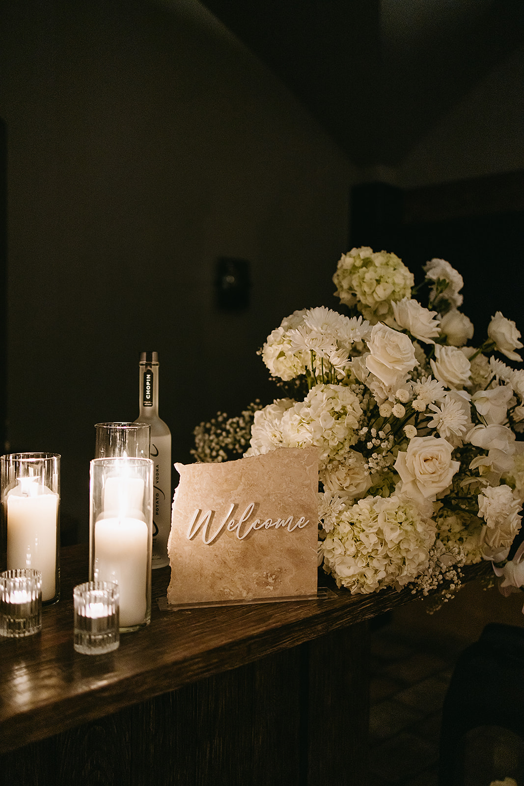 A dimly lit table displays white flowers, candles, and a sign reading "Welcome.