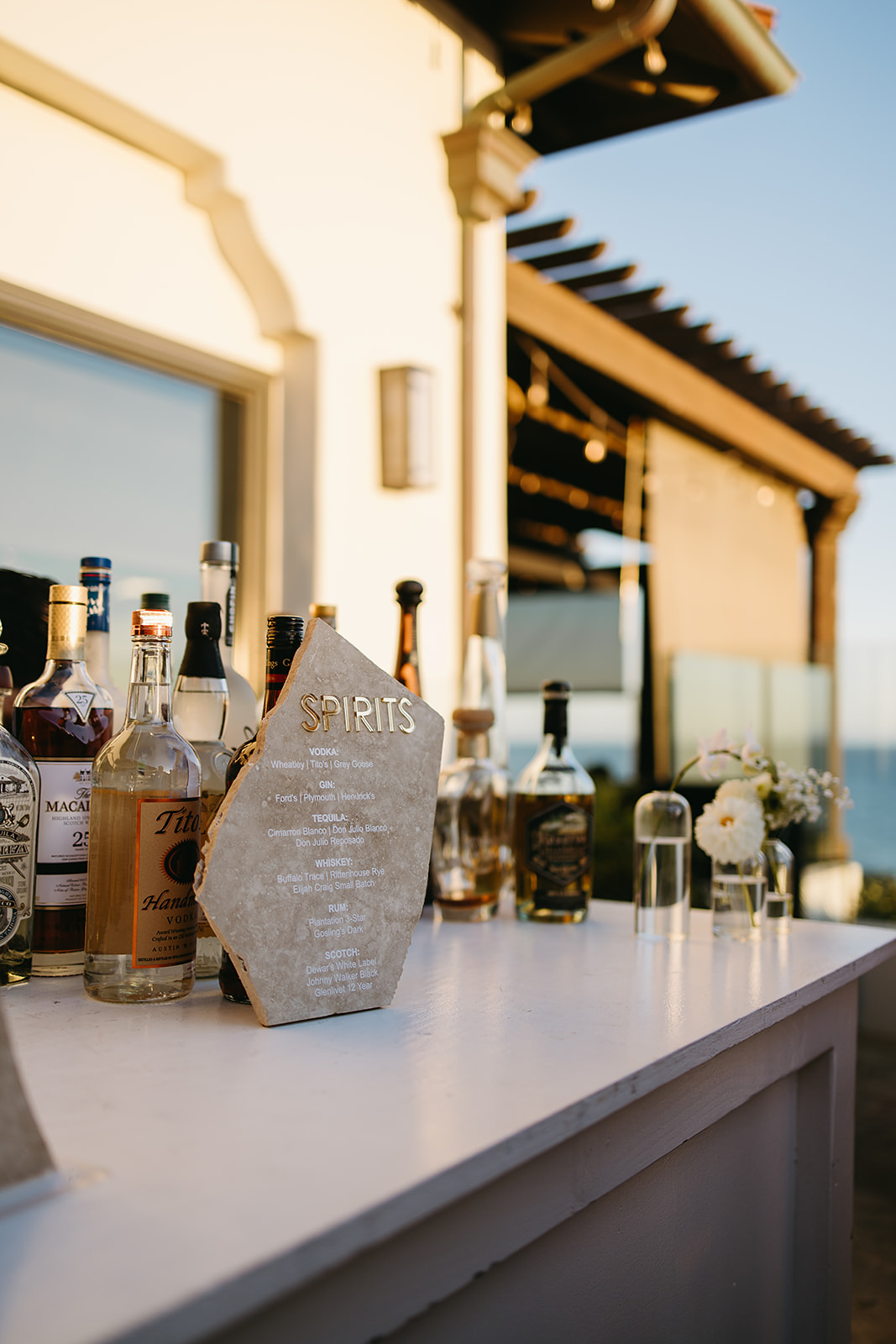 Bottles of various spirits displayed on an outdoor bar counter with a wooden menu on a sunny day.