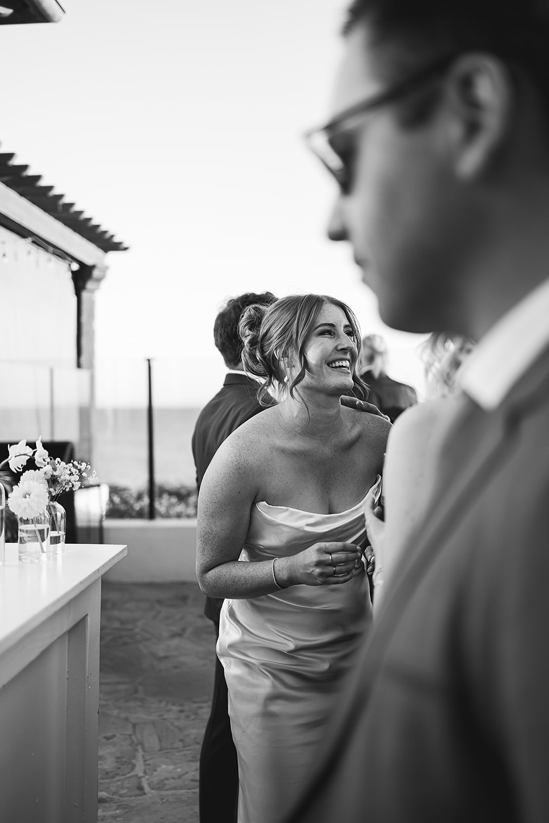 Black and white photo of a woman smiling and holding a drink at an outdoor event, with people and a counter in the background.