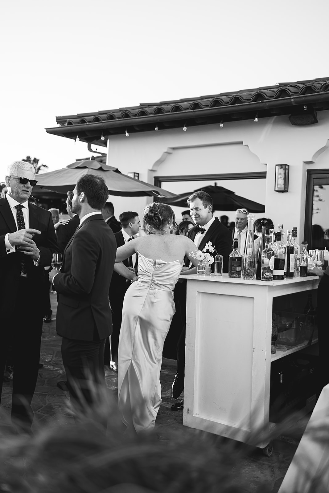  A black and white photo of people socializing at an outdoor event near a bar. Several individuals are dressed formally, and the setting includes umbrellas and a building in the background.