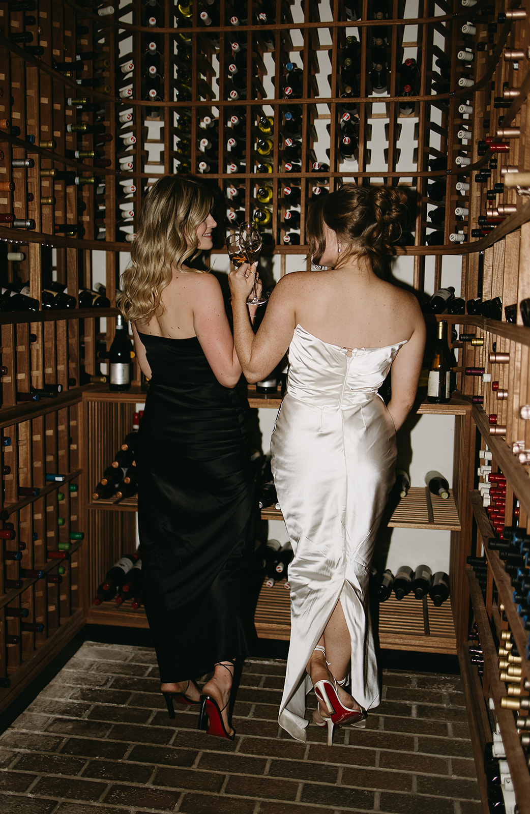 Two women in elegant dresses stand in a wine cellar, facing away, holding a glass of wine each. Shelves filled with wine bottles surround them.