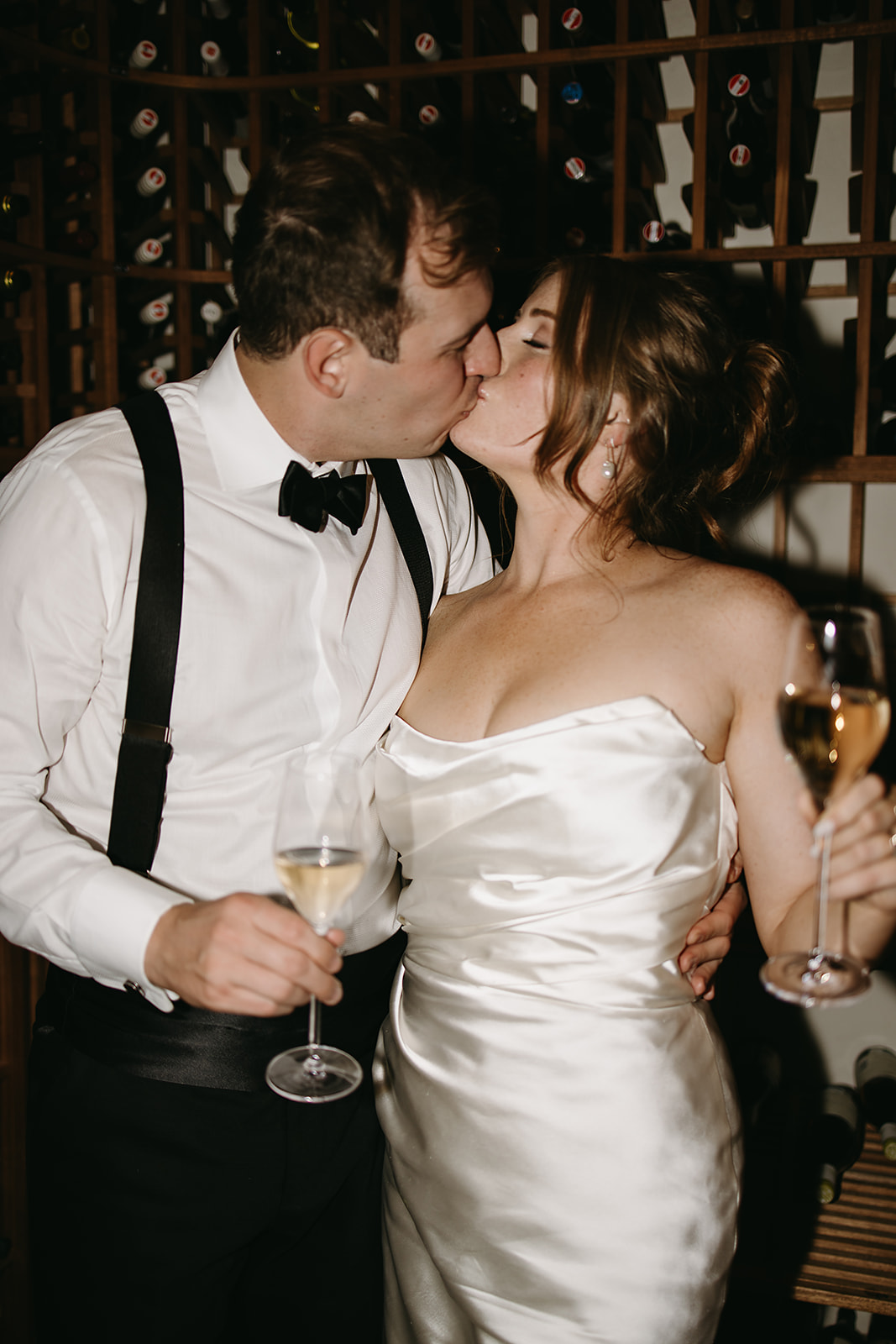 A couple in formal attire shares a kiss while holding wine glasses in front of a wine rack.