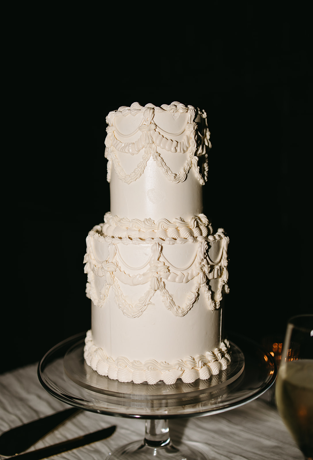 A two-tier white cake with decorative frosting swirls on a glass stand, placed on a table with a glass of liquid nearby.