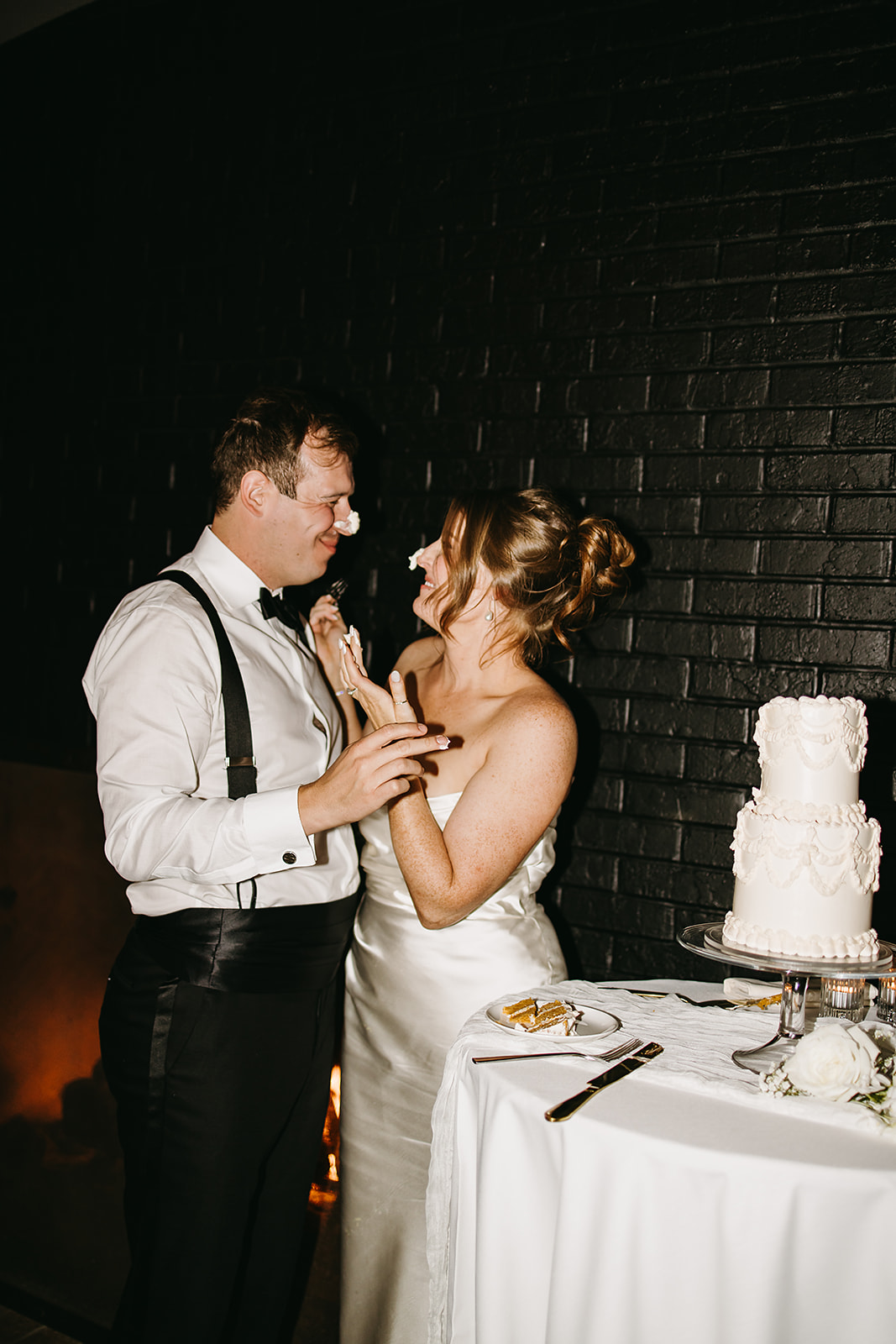 A couple stands by a table with a cake. The woman playfully puts frosting on the man's nose. Both are dressed in formal attire. 