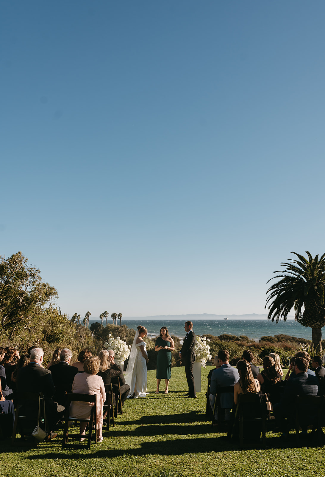 Outdoor wedding ceremony with guests seated on a lawn facing a couple and officiant, near the ocean with palm trees in the background under a clear blue sky at Ritz Carlton Bacara in Santa Barbara 