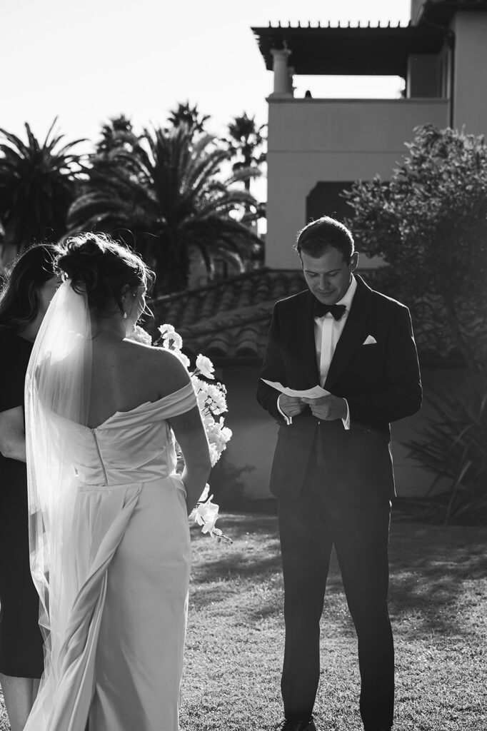 Outdoor wedding ceremony with guests seated on a lawn facing a couple and officiant, near the ocean with palm trees in the background under a clear blue sky at Ritz Carlton Bacara in Santa Barbara 