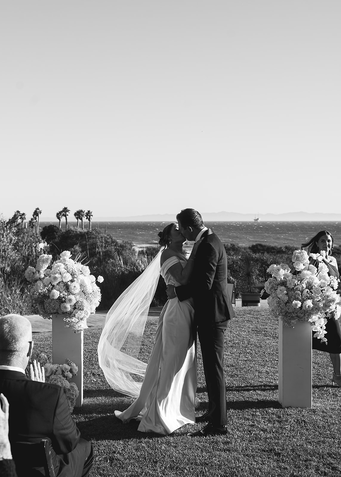 Outdoor wedding ceremony with guests seated on a lawn facing a couple and officiant, near the ocean with palm trees in the background under a clear blue sky at Ritz Carlton Bacara in Santa Barbara 