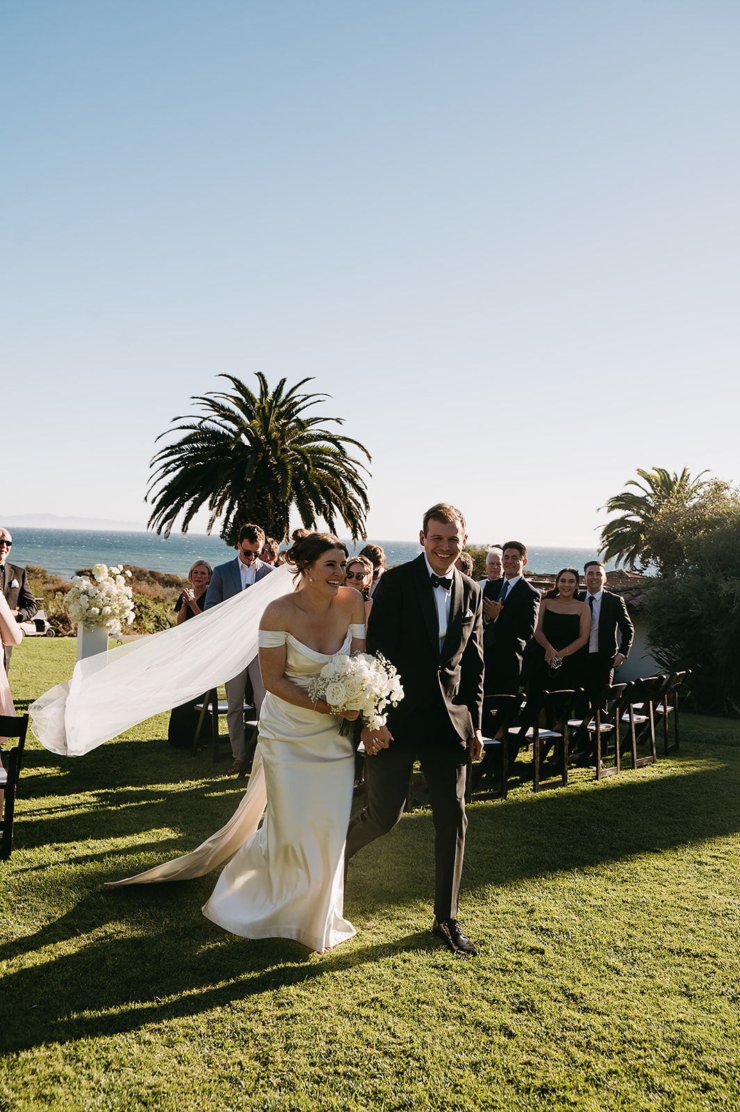 Outdoor wedding ceremony with guests seated on a lawn facing a couple and officiant, near the ocean with palm trees in the background under a clear blue sky at Ritz Carlton Bacara in Santa Barbara 