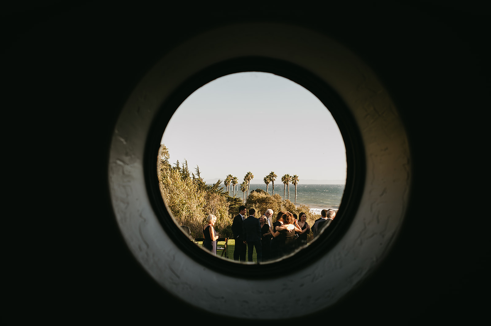 A group of people gathered outside near a palm tree, viewed through an archway. Ocean visible in the background. 