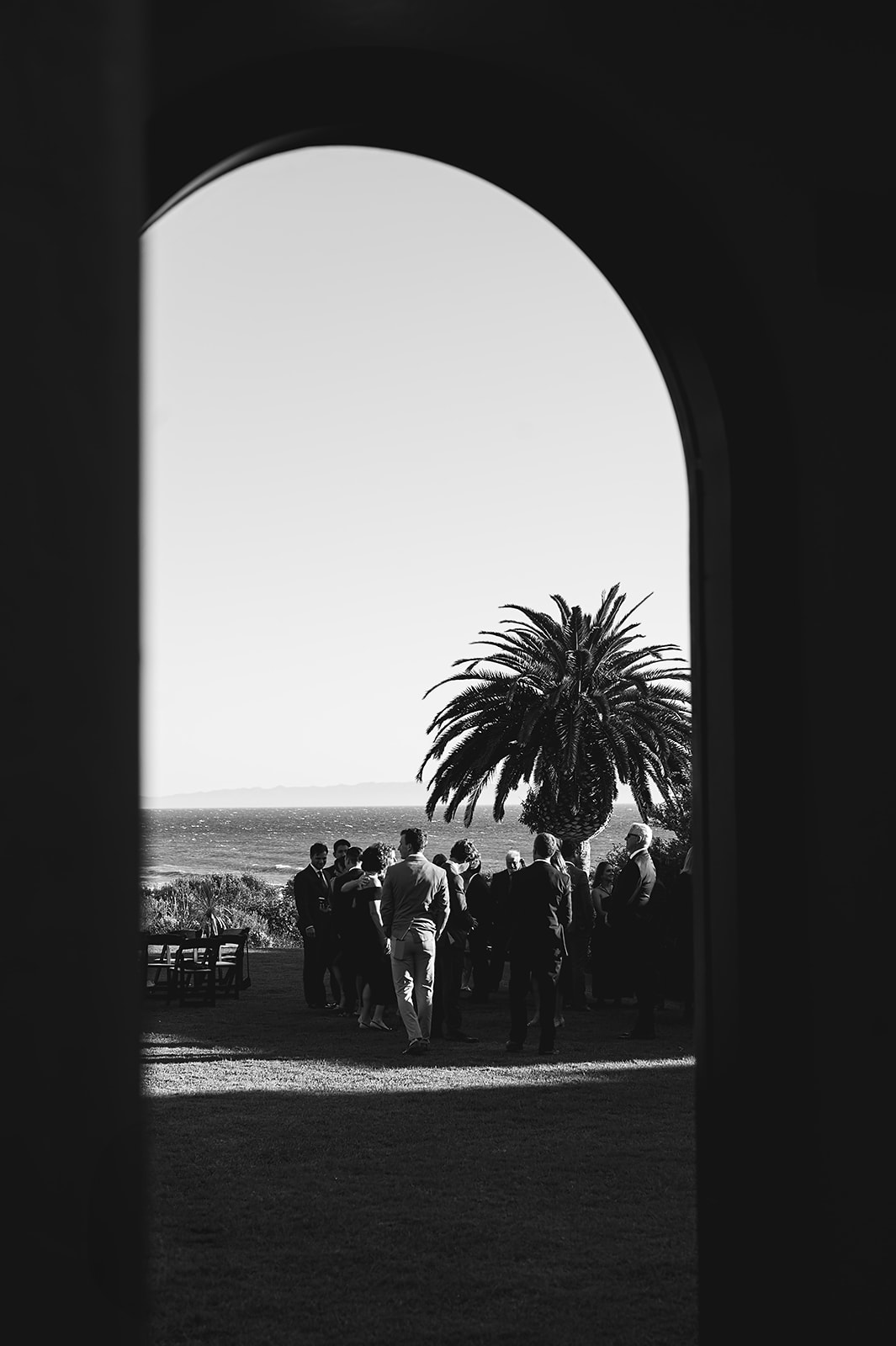 A group of people gathered outside near a palm tree, viewed through an archway. Ocean visible in the background. Black and white image.