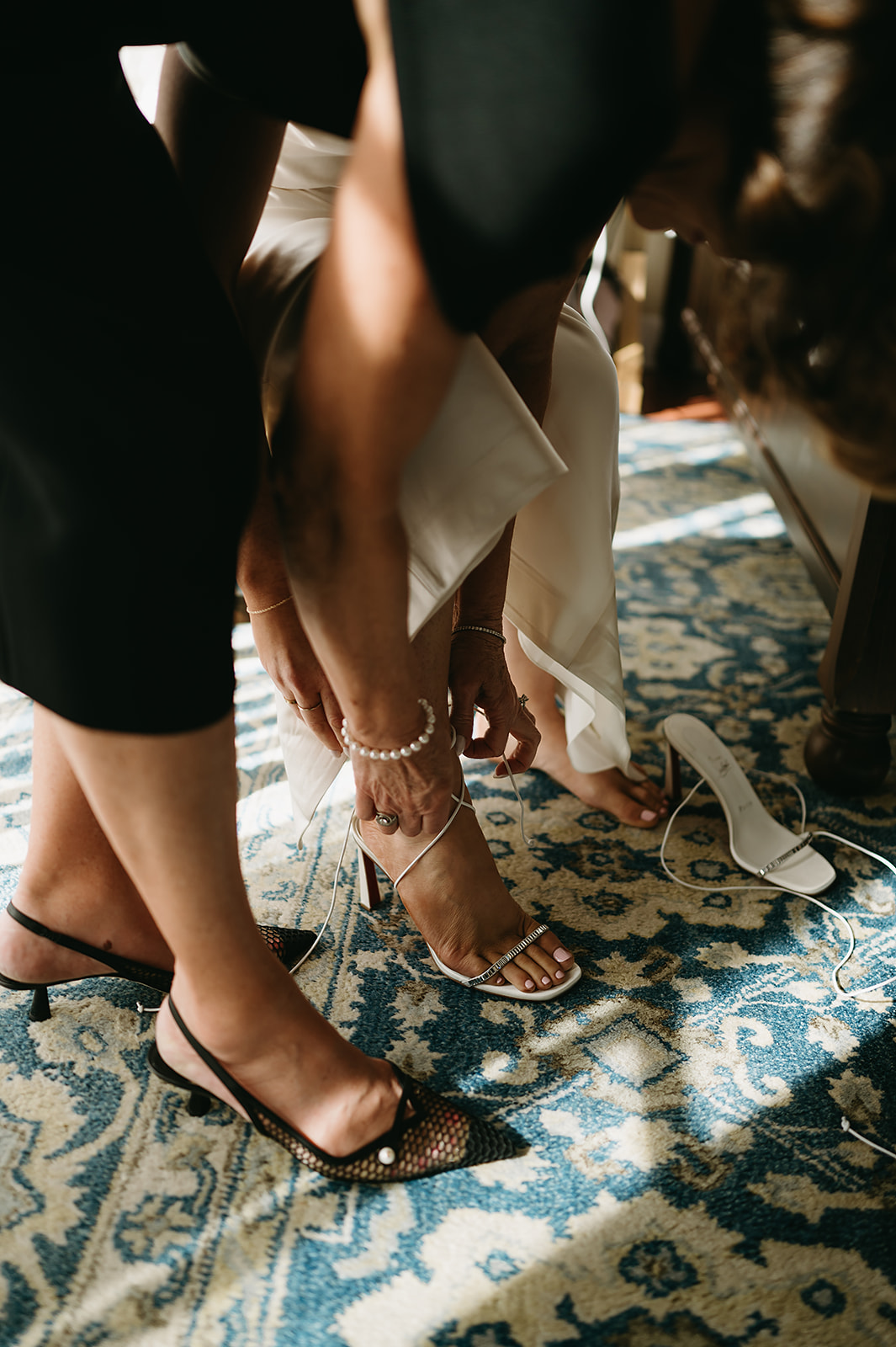 Two people adjusting a woman's high-heeled shoe strap over a patterned carpet.