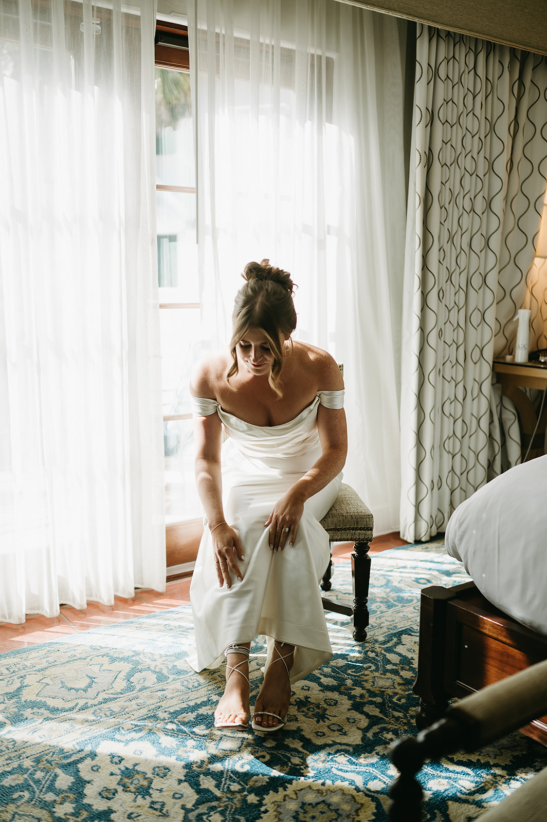 A woman in a white off-shoulder dress sits on a chair in a brightly lit room with patterned carpet and curtains.