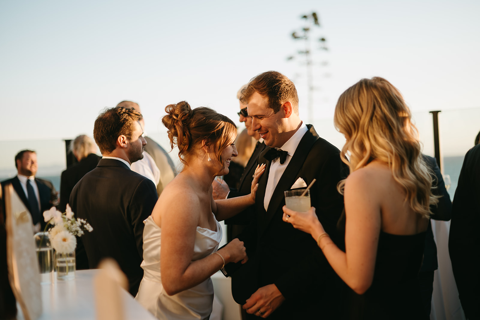 People in formal attire mingling at an event outdoors, with a clear sky in the background.