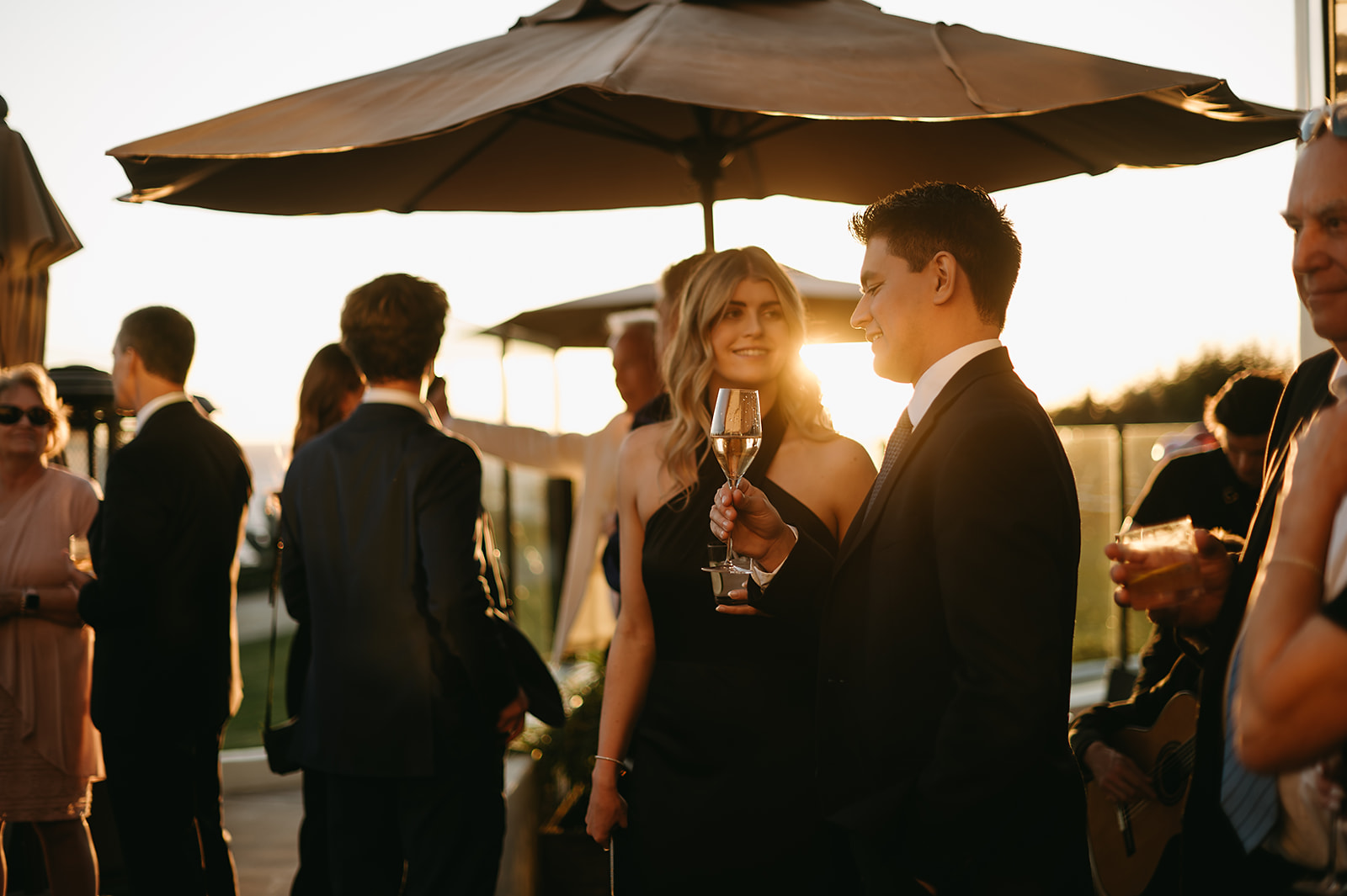 People in formal attire mingling at an event outdoors, with a clear sky in the background.