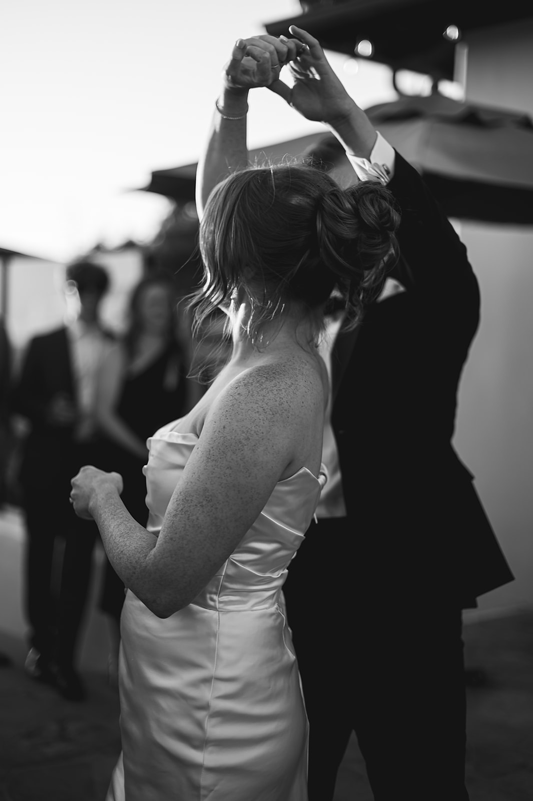 A couple dances together outdoors at a wedding. The groom is in a tuxedo, and the bride is in a white dress. Two men in suits stand in the background.