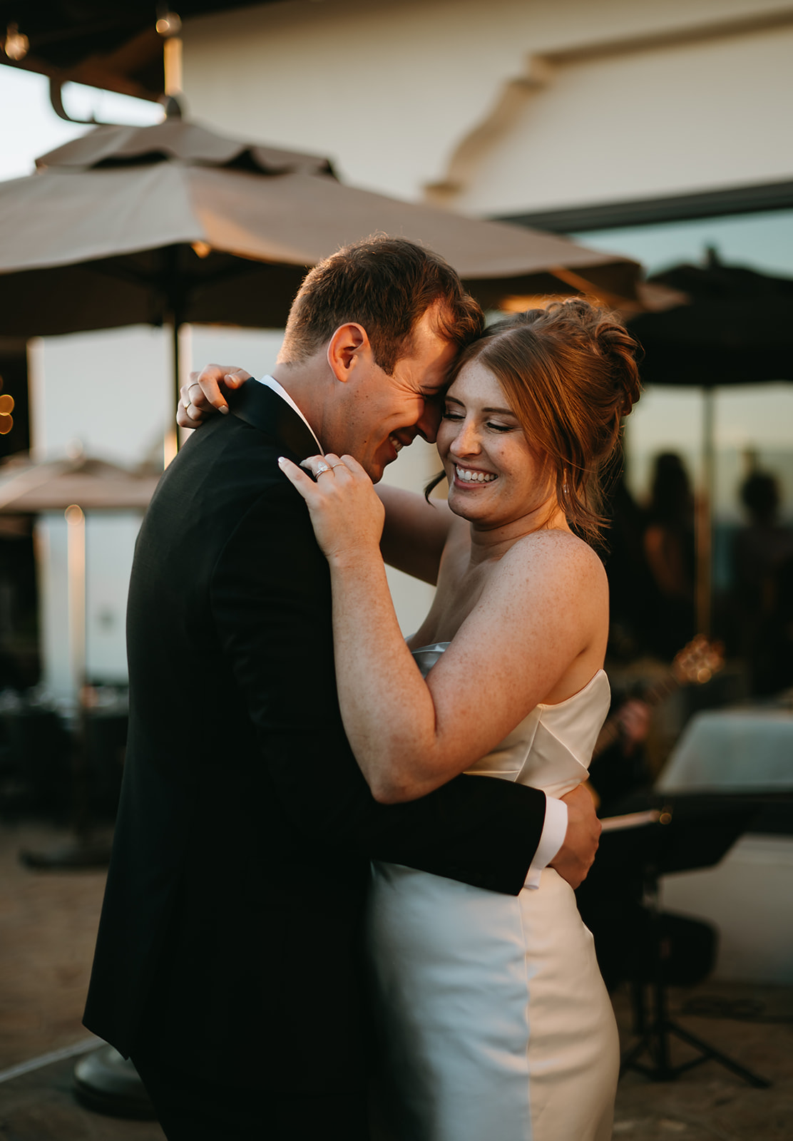 A couple dances together outdoors at a wedding. The groom is in a tuxedo, and the bride is in a white dress. Two men in suits stand in the background.