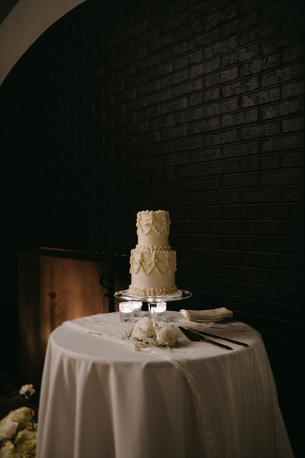 A two-tier white cake with textured icing sits on a round, draped table, accompanied by a cake knife and spatula, in a dimly lit room with dark brick walls.