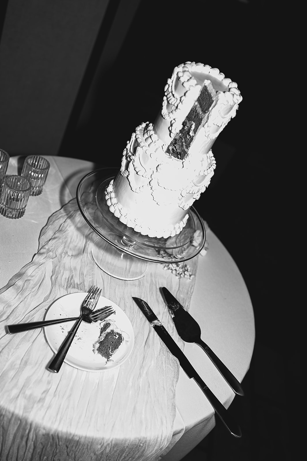 A two-tier white cake with textured icing sits on a round, draped table, accompanied by a cake knife and spatula, in a dimly lit room with dark brick walls.