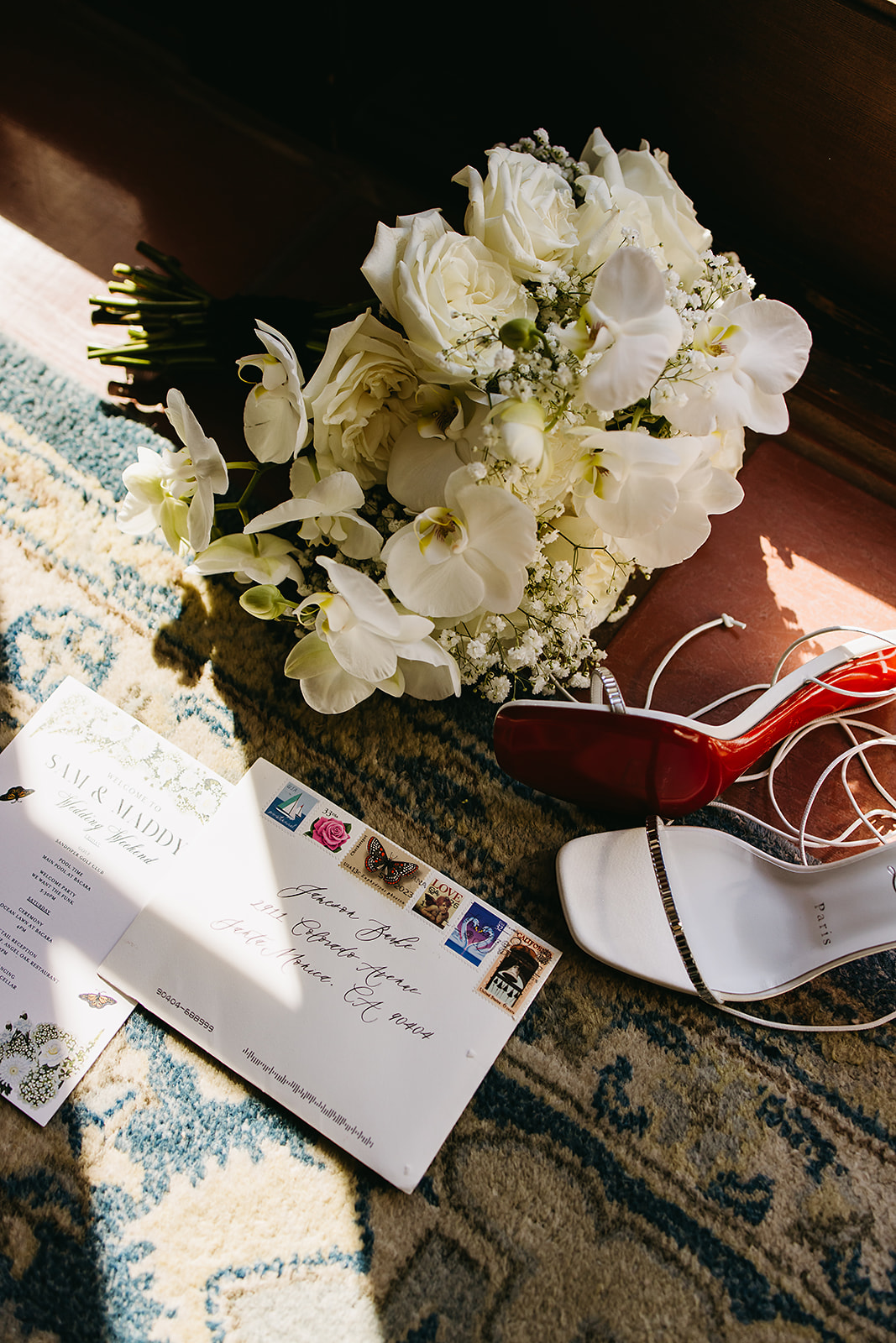 A bouquet of white flowers, white high-heeled shoes, and an envelope with stamps are placed on a patterned rug.