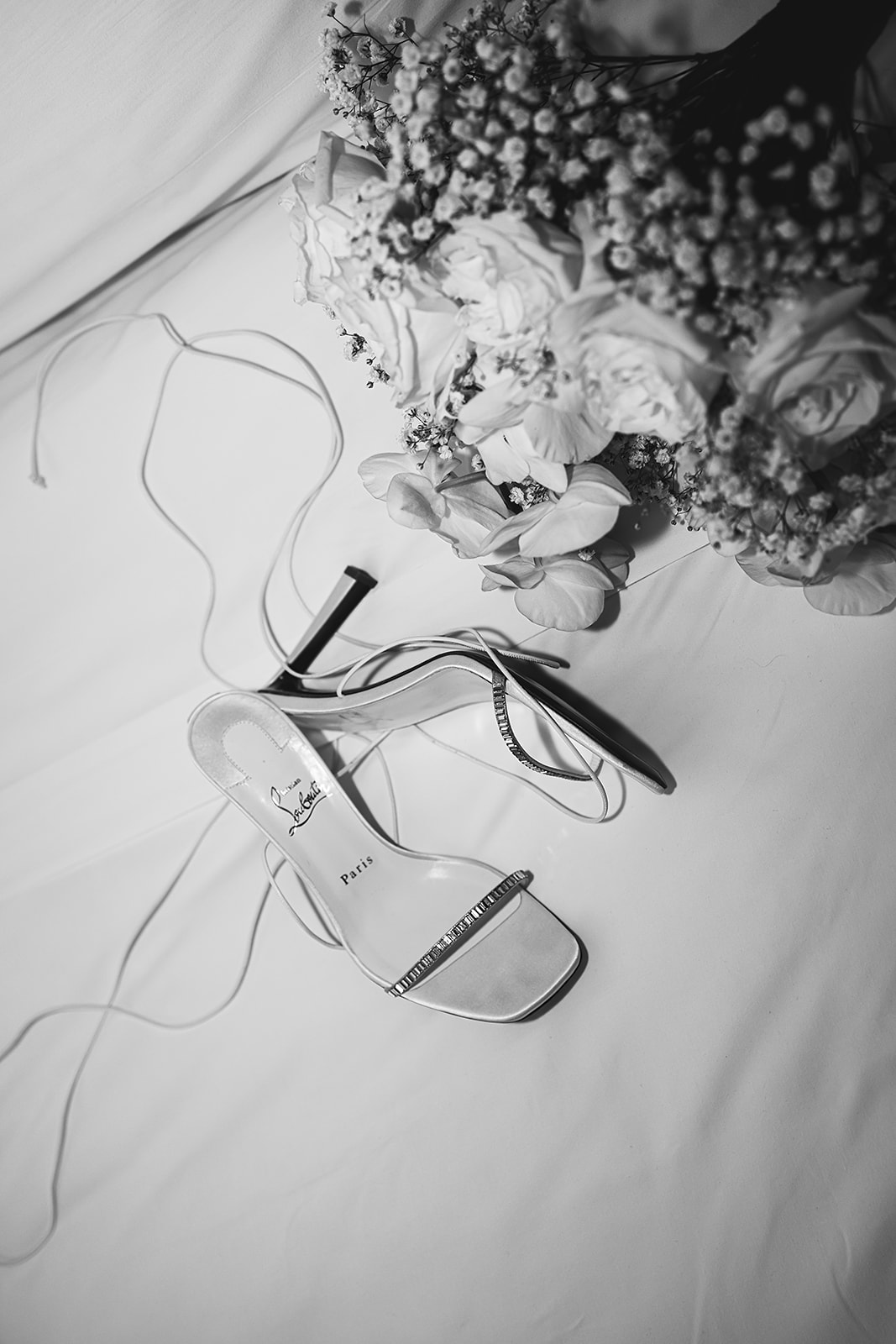 A bouquet of white flowers, white high-heeled shoes, and an envelope with stamps are placed on a patterned rug.