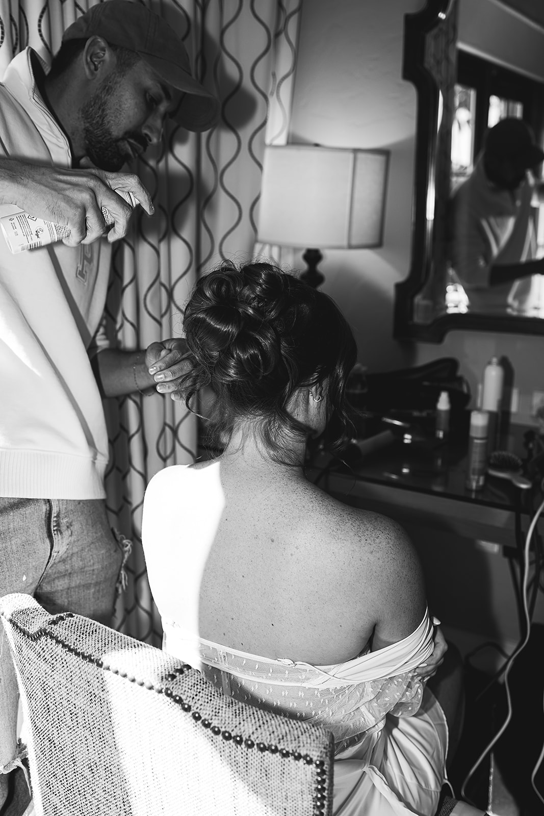 A man styles a woman's hair in front of a vanity mirror. The woman is seated and wears an off-shoulder top. Various hair products are on the vanity.