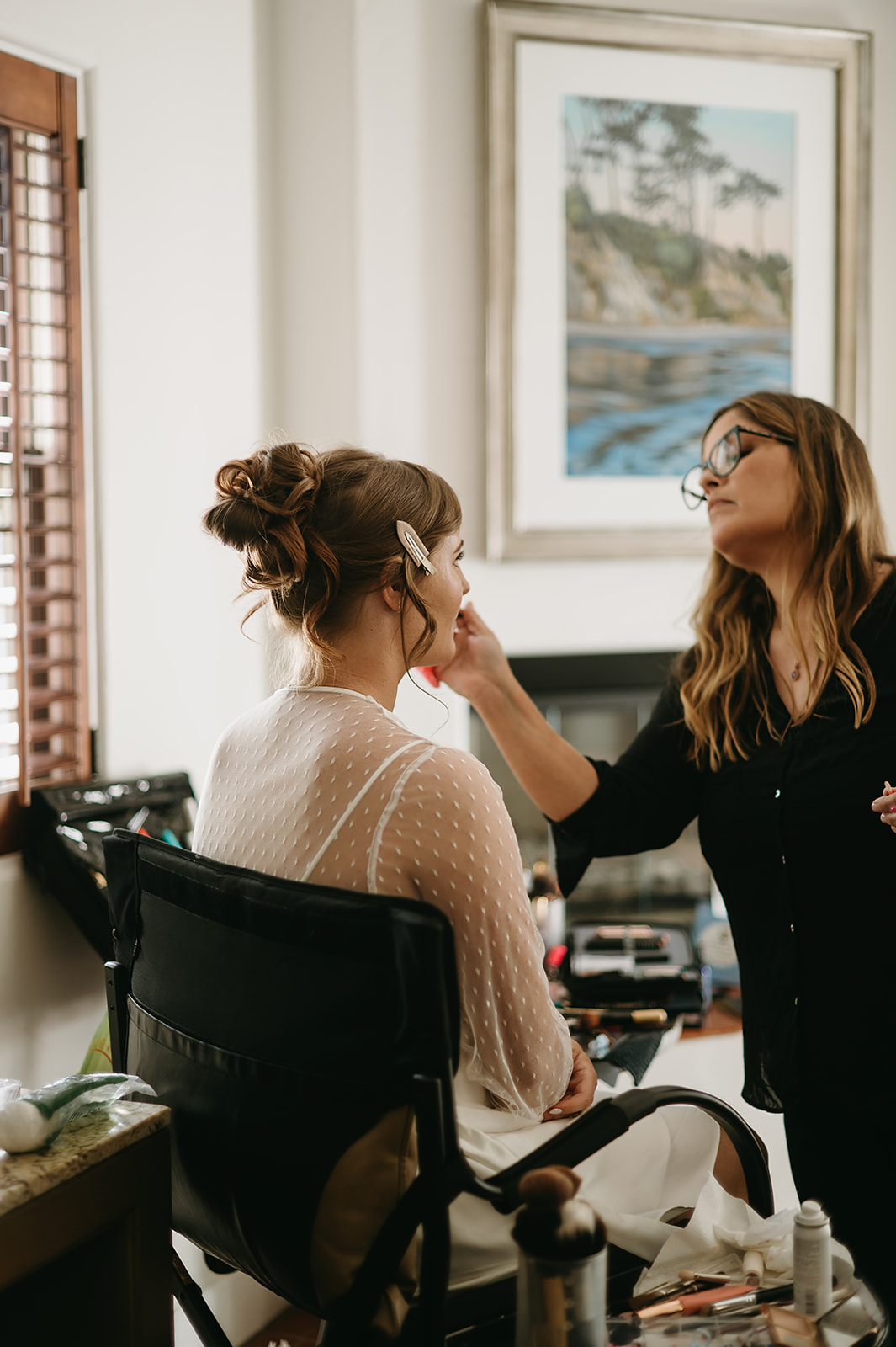 A woman is sitting on a chair while a makeup artist applies makeup to her face in a well-lit room.