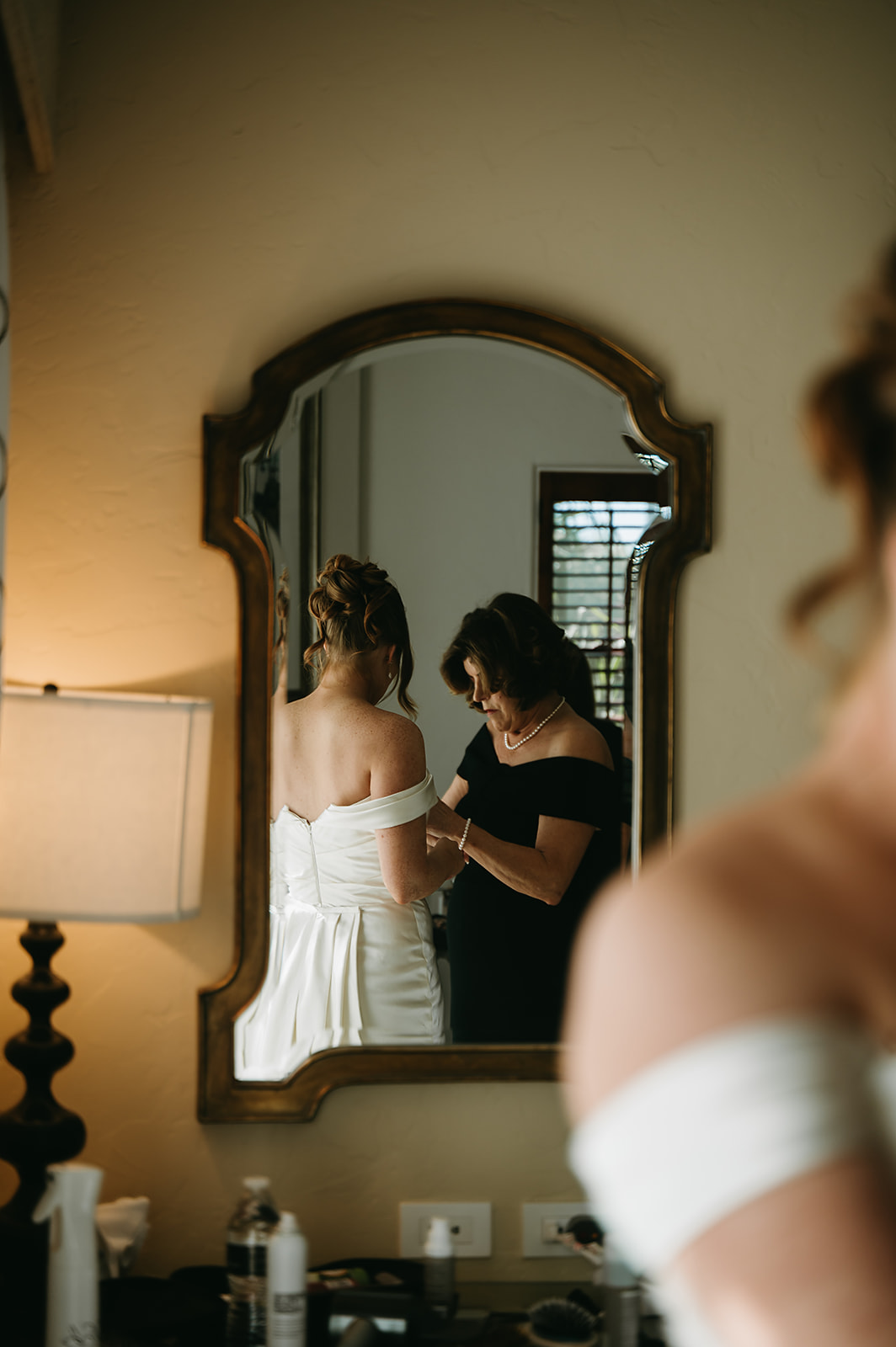 A woman in a white dress is being helped by another woman, reflected in a wall mirror.