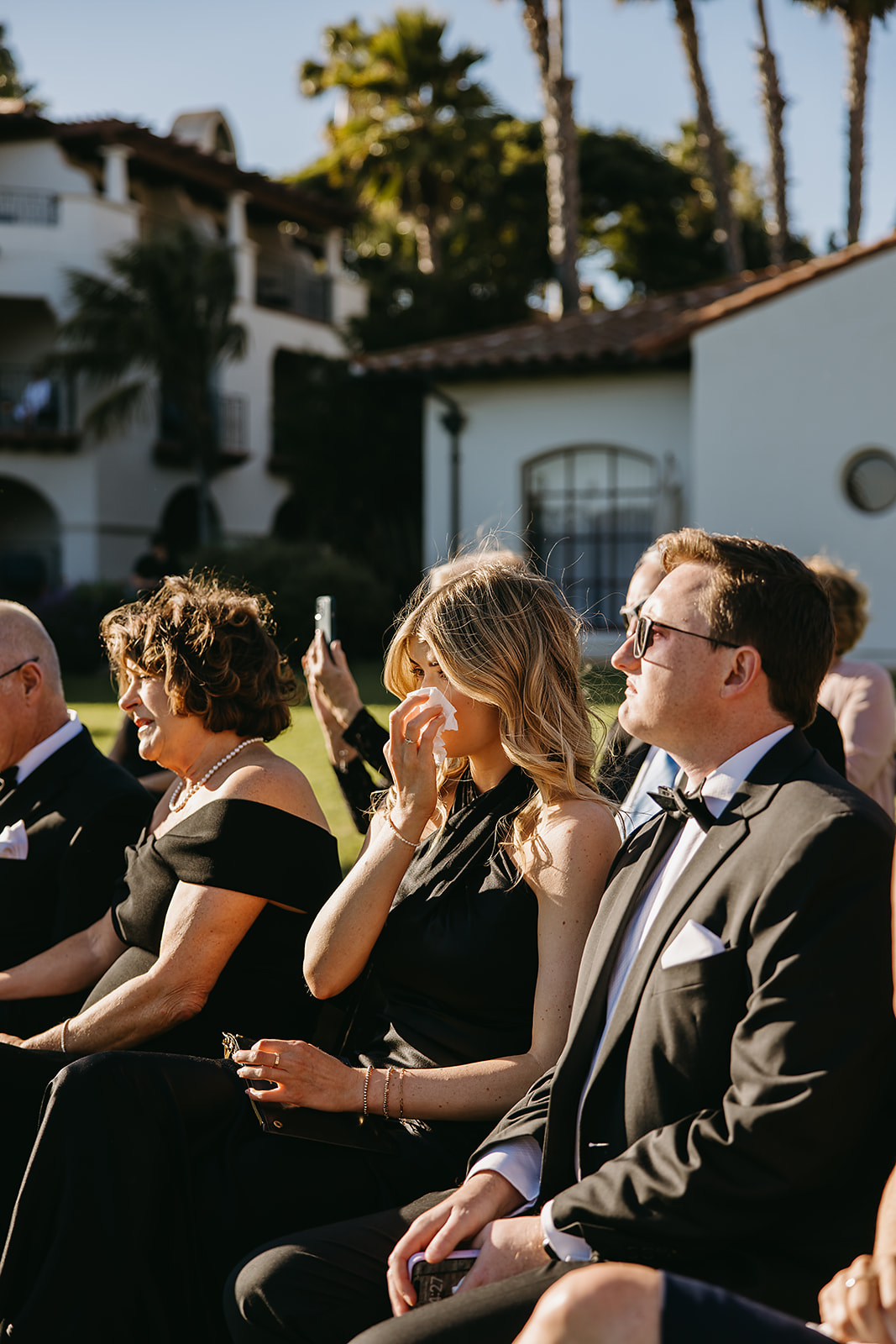 People seated outdoors at an event, dressed formally. One woman wipes her eye, while others look forward. Sunlight and palm trees in the background.