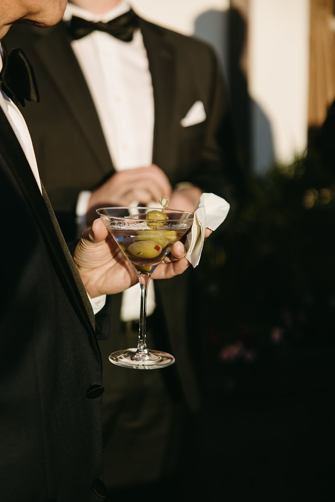 Two people in tuxedos, one holding a martini with olives and a napkin.