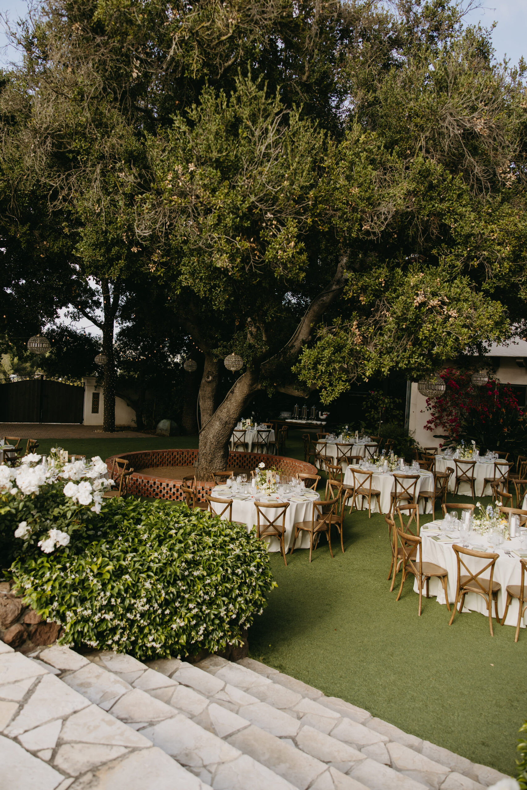 Outdoor dining area with round tables covered in white tablecloths. Wooden chairs surround the tables. Overhead, chandeliers hang from tree branches. Lush greenery is visible in the background at quail ranch