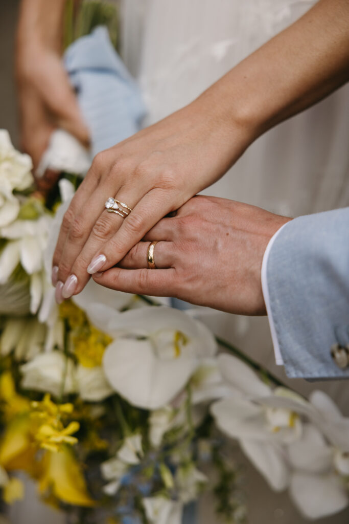 Two hands with wedding rings rest on a bouquet of white and yellow flowers.