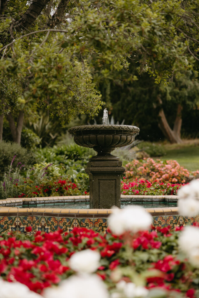 A stone fountain in a garden surrounded by red and white flowers, with trees in the background.