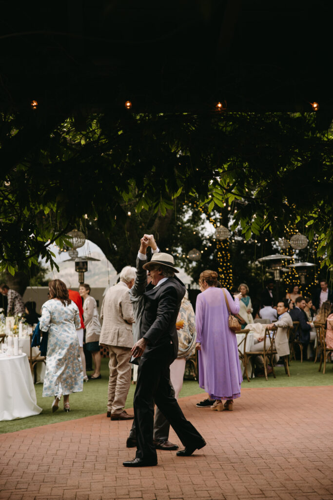 A couple dancing outdoors at a wedding reception. Guests in formal attire are seated at tables under trees with string lights.