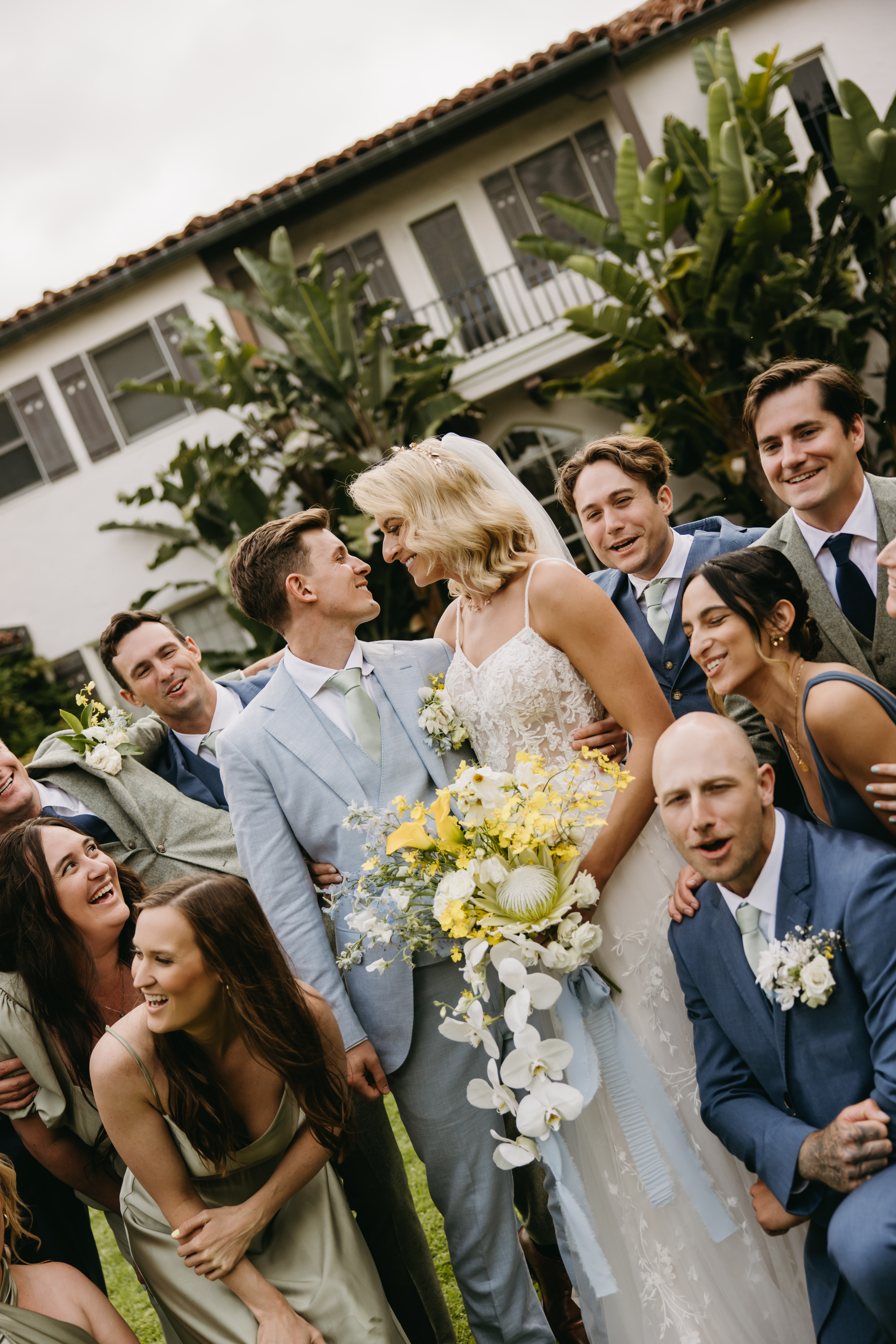 A wedding couple in light attire stands with a group of smiling people outdoors. The bride holds a bouquet with yellow flowers, and a building with large windows is in the background.