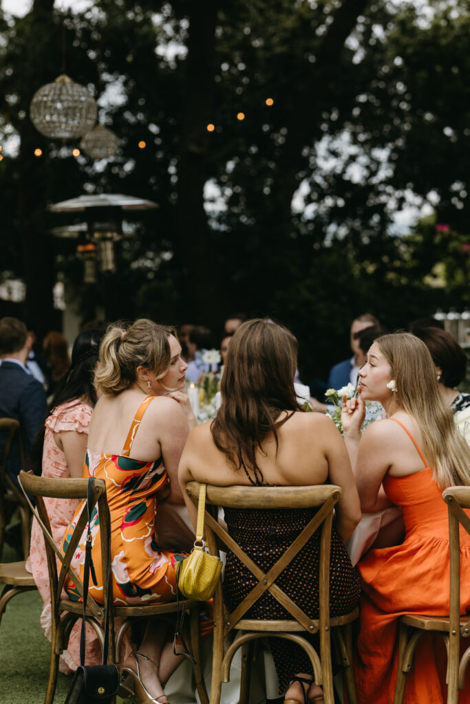 Outdoor gathering with people seated on wooden chairs at a table. Three women in colorful dresses are seen from the back, surrounded by greenery and string lights a quail ranch