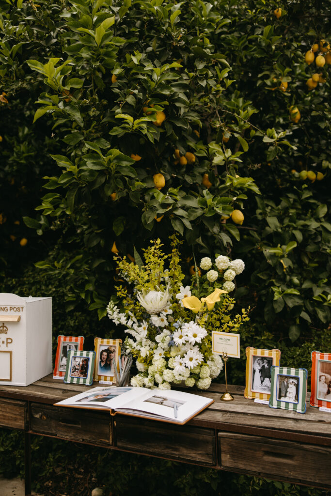 A table with an open book, framed photos, a floral arrangement, and a box, set against a background of lush green foliage with yellow fruits.